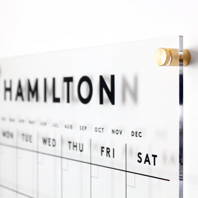 Close-up of a modern acrylic wall calendar with the word HAMILTON at the top. Days of the week, Monday to Saturday, are visible in bold black lettering. Gold mounting hardware is visible on the right side.