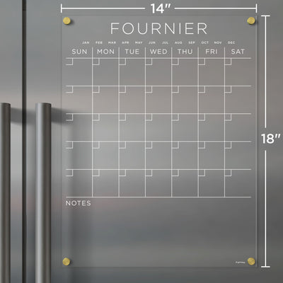 The Girl Friday Acrylic Fridge Calendar, customizable with a family name, features a clear acrylic design with gold hardware, an empty days and months grid, a NOTES section at the bottom, and includes wet-erase markers. It measures 14 inches wide by 18 inches tall.