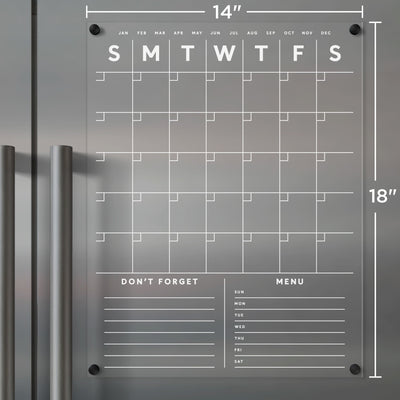 A Clear Acrylic Monthly Fridge Calendar by Girl Friday Shop, perfect for wet-erase markers, is mounted on a gray wall. It features month and weekday headers, customizable sections for Dont Forget and Menu, ample note space, black corner magnets, and silver handles.