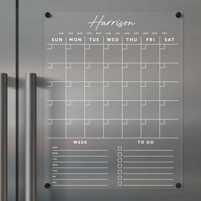 The Girl Friday Clear Acrylic Monthly Fridge Calendar beautifully showcases a custom family name, Harrison, at the top. Mounted on a wall, it features a blank monthly grid and customizable sections for Week and To Do, allowing notes to be easily written with wet-erase markers.
