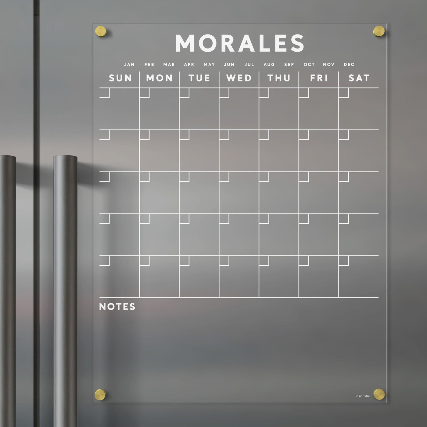The Acrylic Fridge Calendar by Girl Friday, customizable with your family name in white text, is mounted on a gray surface with silver handles. It displays days and months at the top and includes a NOTES section at the bottom. Use wet-erase markers to keep your weekly plans organized.