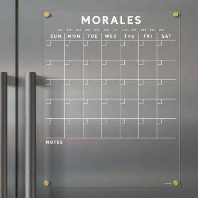 The Acrylic Fridge Calendar by Girl Friday, customizable with your family name in white text, is mounted on a gray surface with silver handles. It displays days and months at the top and includes a NOTES section at the bottom. Use wet-erase markers to keep your weekly plans organized.