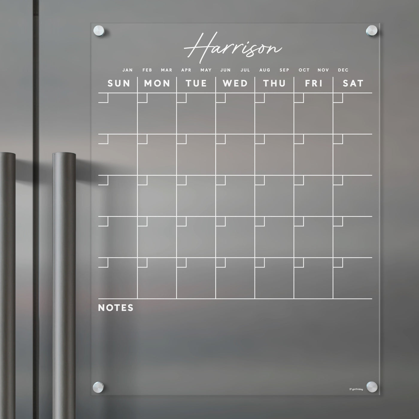 A Harrison clear acrylic calendar by Girl Friday is mounted on a dark surface with two handles. Its ideal for wet-erase markers and features days, months, and a bottom notes section.