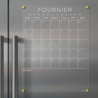 Girl Fridays Acrylic Fridge Calendar, customizable with a family name, spans January to December and features a Sunday-to-Saturday layout with a notes section. Designed for use with wet-erase markers, it easily mounts on metallic surfaces.