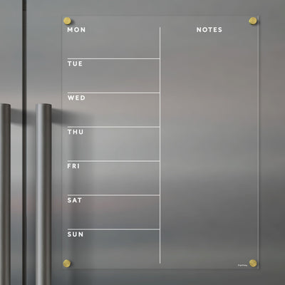 The Girl Friday Clear Acrylic Fridge Calendar is a minimalistic glass weekly planner featuring Mon to Sun columns and Notes. It includes gold corner pegs, two gray handles on the left, and is compatible with wet-erase markers for easy updates.