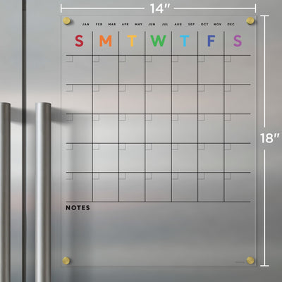 The Clear Acrylic Fridge Calendar with Rainbow Text by Girl Friday Shop attaches to a metal surface and is ideal for dry-erase markers. It has a weekly layout with colorful initials for days, monthly headers, note space at the bottom, and measures 14 x 18.