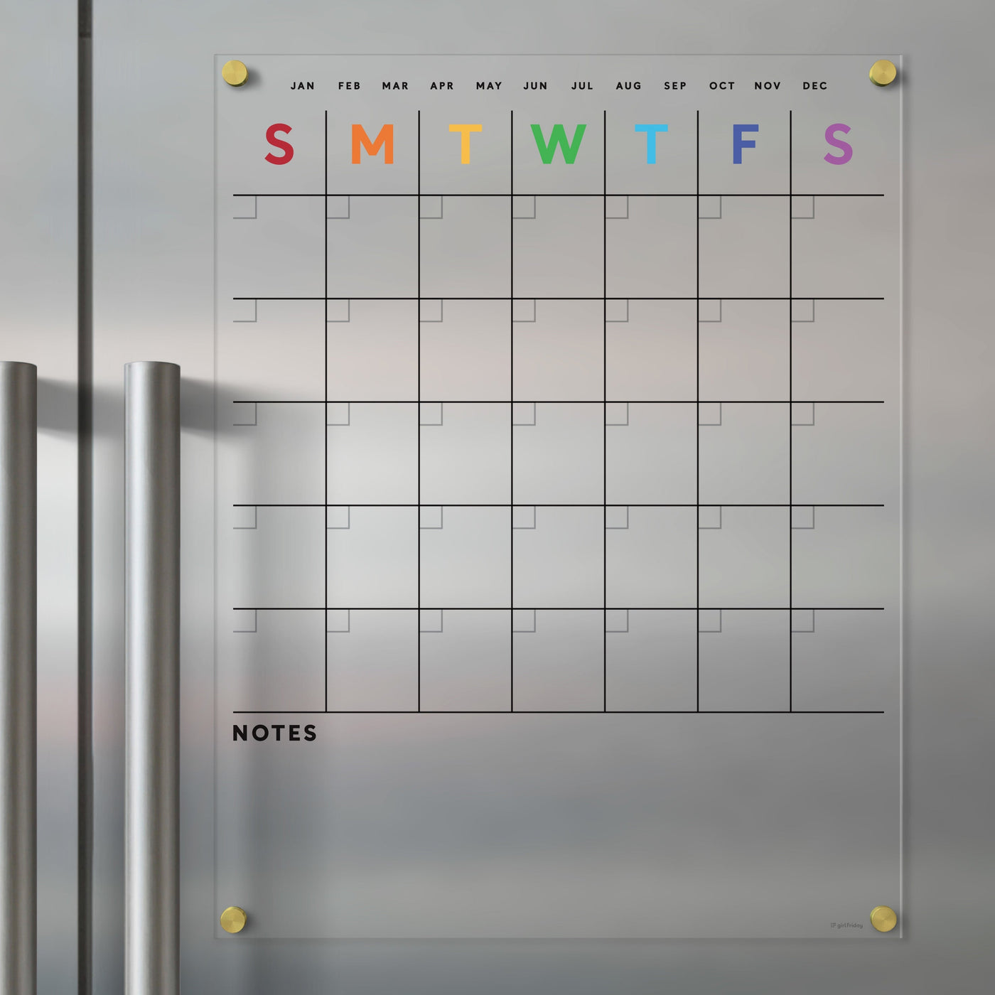The Clear Acrylic Fridge Calendar from Girl Friday Shop displays a rainbow-colored grid for the week against a metallic surface. Months are listed in black above, with a notes section below. Stainless steel magnets keep it all organized.