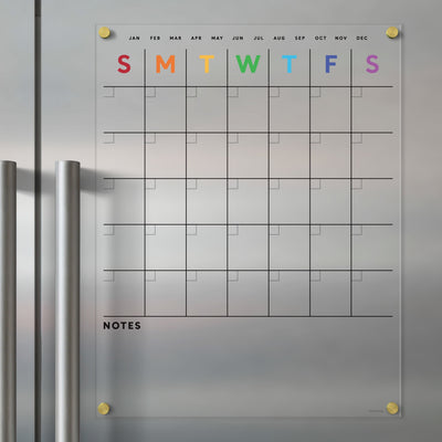 The Clear Acrylic Fridge Calendar from Girl Friday Shop displays a rainbow-colored grid for the week against a metallic surface. Months are listed in black above, with a notes section below. Stainless steel magnets keep it all organized.