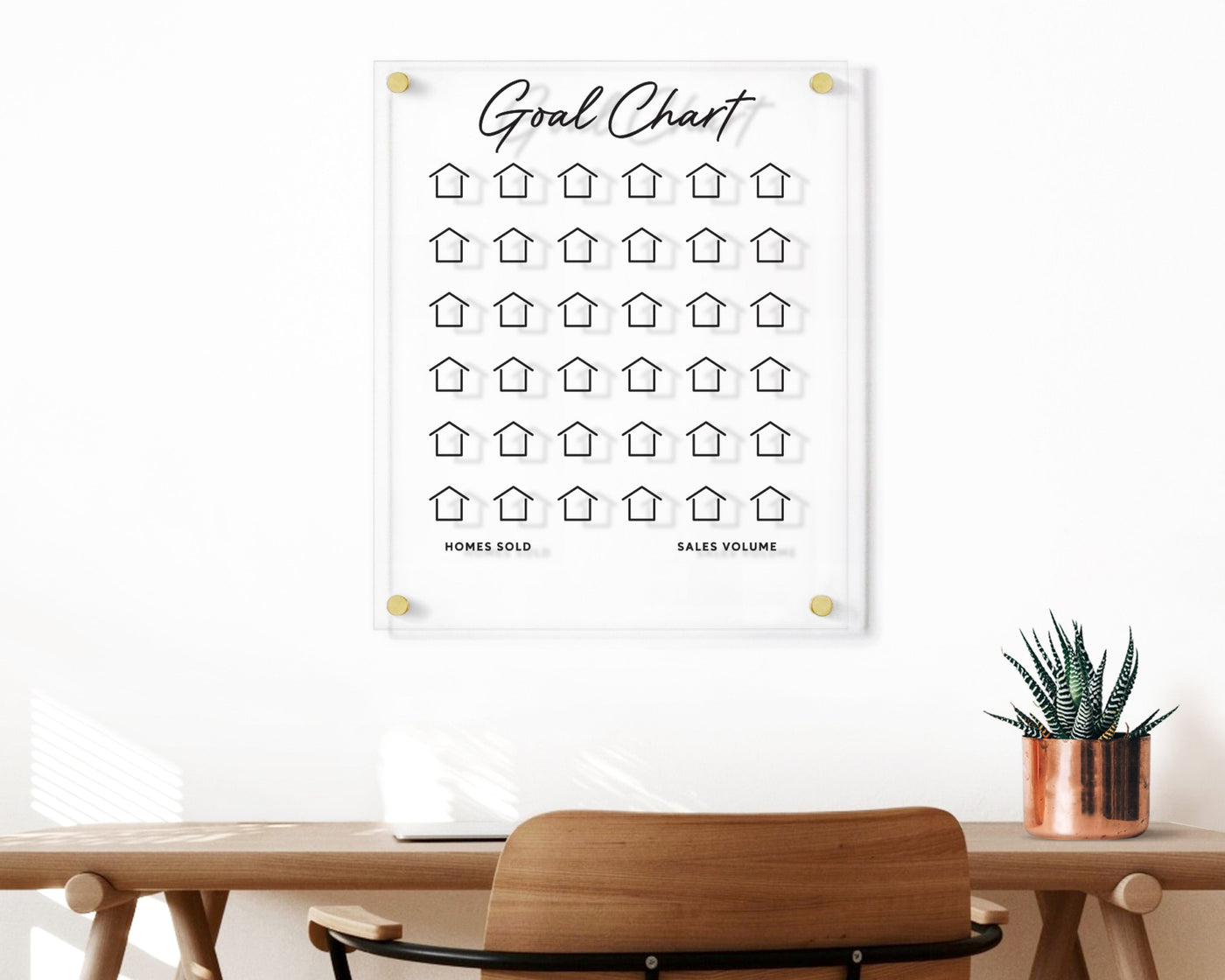 The Clear Acrylic Goal Chart by Girl Friday features a 5x6 grid of house icons with Homes Sold and Sales Volume text below. A wooden desk, complete with a chair, colored markers, and a potted plant, sits in the foreground for an organized workspace.