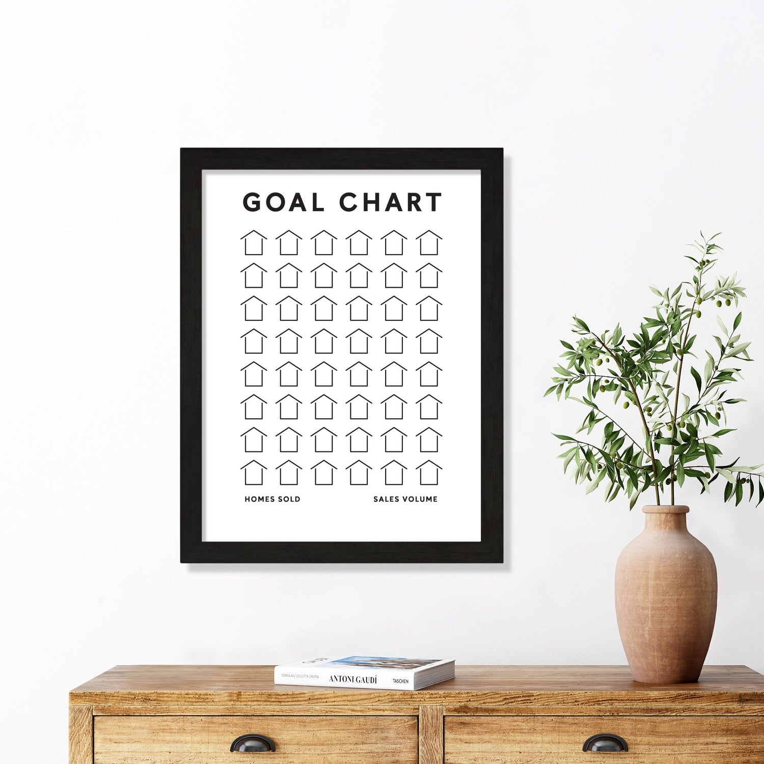 A Goal Chart Poster Print by Girl Friday Shop elegantly decorates a white wall above a wooden table. The chart, featuring rows of outlined house icons, is complemented by a vase with green branches and two books, blending functionality and style seamlessly.