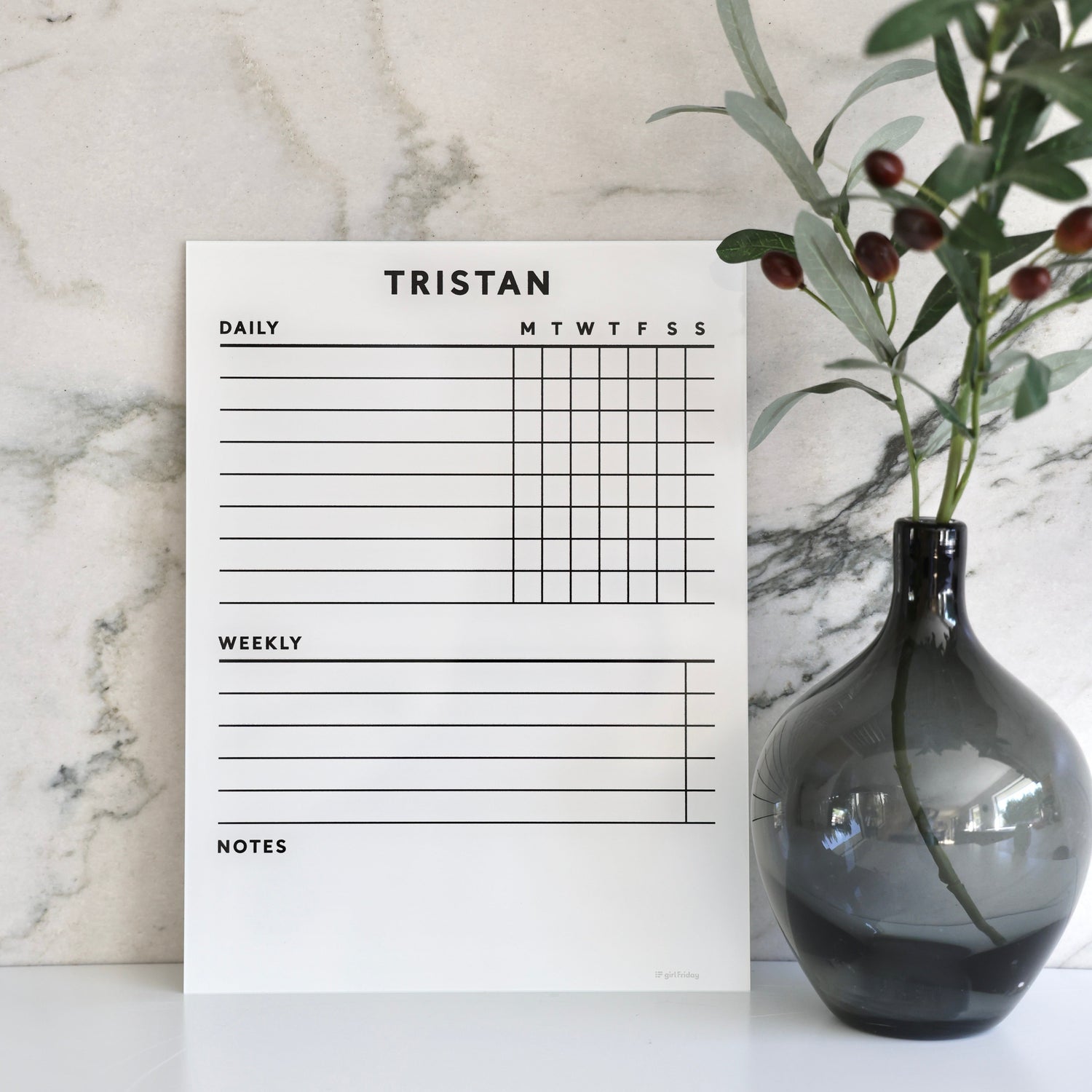 On a marble background, the White Acrylic Chore Chart by Girl Friday Shop showcases daily columns. The name Tristan is styled at the top. Nearby, a dark glass vase with green leaves and small berries enhances the scene. Its customizable with a premium dry erase surface, ideal for non-magnetic fridges.