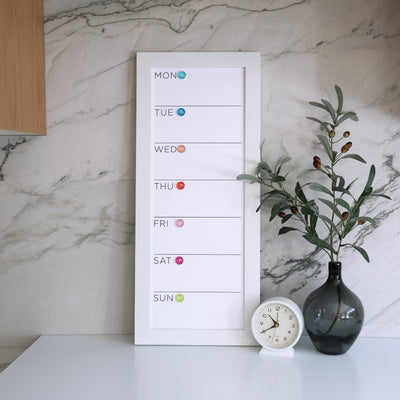 The CalendarGirlCompanys 9 x 24 weekly whiteboard calendar (#923) is mounted on a marble wall. It features a vertical Monday-to-Sunday schedule with colored circles for each day, complemented by a round clock and a vase with green branches on the premium acrylic surface.