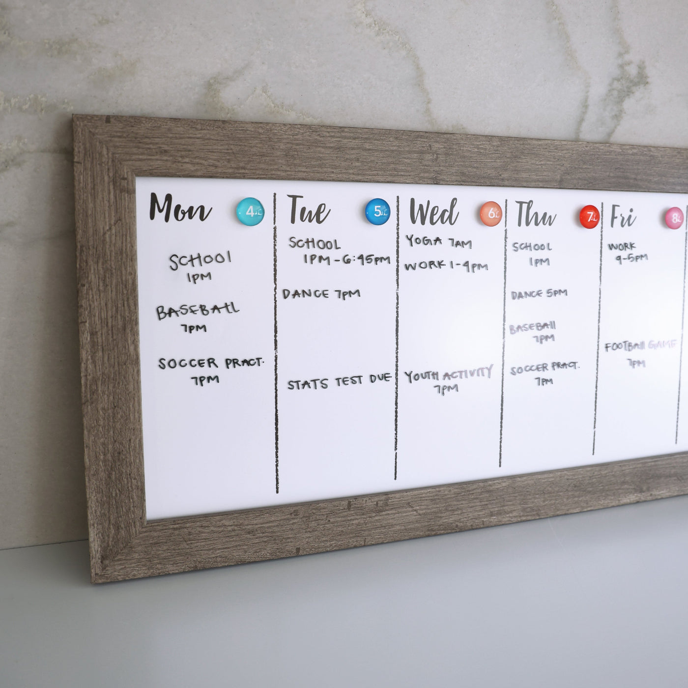 The CalendarGirlCompany 24 X 9 Framed Weekly Whiteboard Calendar (#922) features a premium acrylic surface for vibrant chalk ink markers. Display school, yoga, baseball, dance, and test schedules with colorful magnets in a wooden frame on a light wall for Monday through Friday.