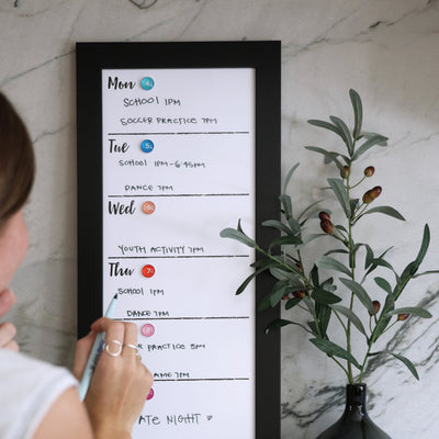 A person uses chalk ink markers on a CalendarGirlCompany 9 X 24 weekly whiteboard calendar (#921) hanging on a marble wall, listing activities like school, soccer practice, and dance. Nearby is an olive branch in a black vase.
