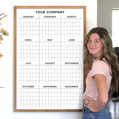 A woman with long hair in a pink shirt stands beside a 2025 Girl Friday Vertical Yearly Calendar titled Your Company, featuring monthly rows. She smiles, hand on hip, with a yellow-flowered plant visible on the left.