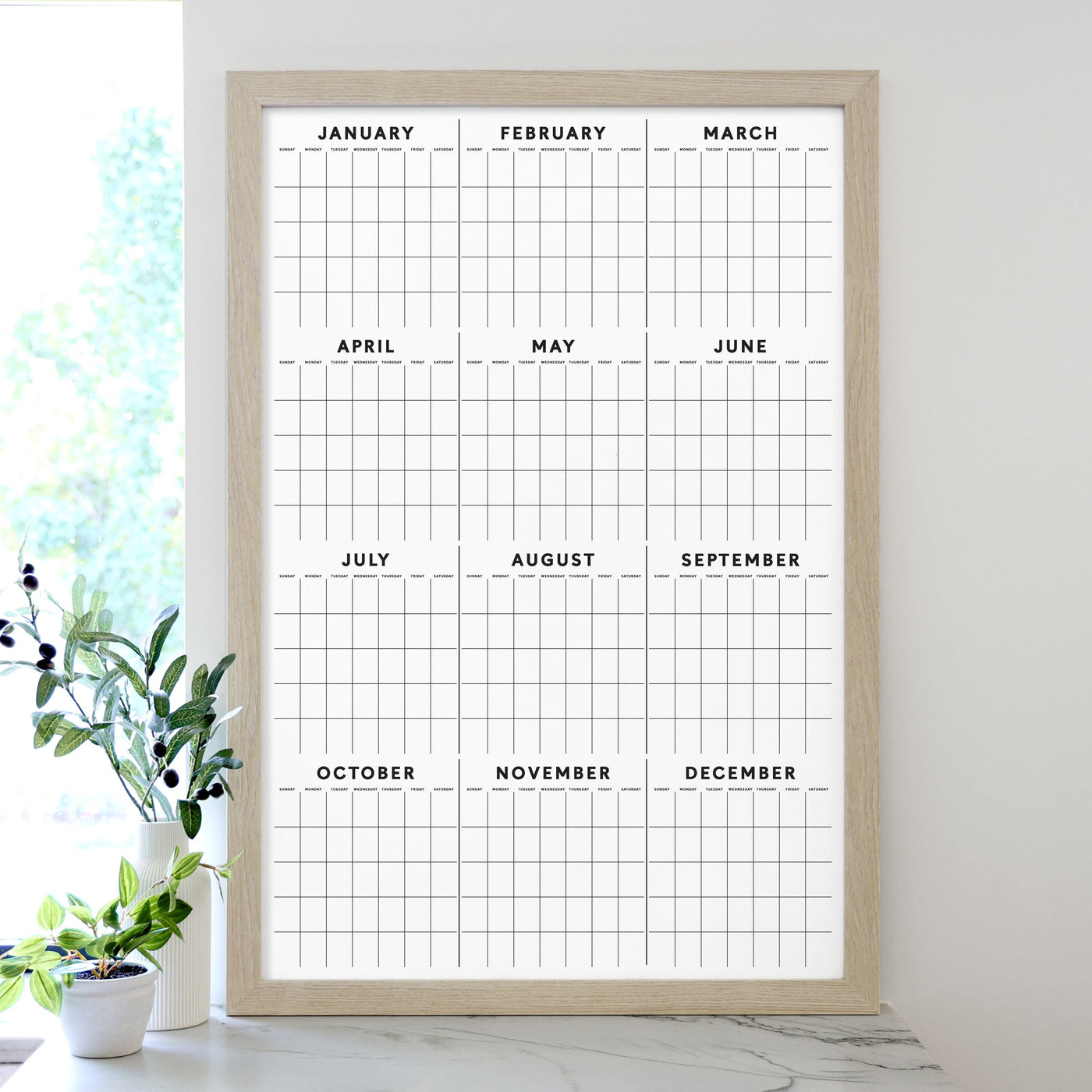 The 2025 Framed Yearly Whiteboard Calendar by Girl Friday features a minimalist design framed in wood, with a premium acrylic surface displaying all months from January to December. Ideal for use with wet-erase markers, its complemented by a small plant sitting nearby on the white surface.