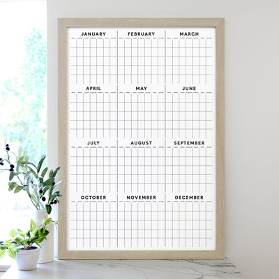 The 2025 Framed Yearly Whiteboard Calendar by Girl Friday features a minimalist design framed in wood, with a premium acrylic surface displaying all months from January to December. Ideal for use with wet-erase markers, its complemented by a small plant sitting nearby on the white surface.