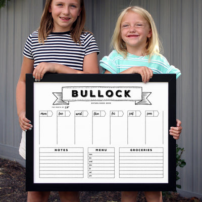 Two kids smile while holding the CalendarGirlCompanys 24 X 18 Framed Whiteboard Calendar, which includes sections for notes, menu, groceries, and a weekly schedule. Theyre in front of a gray ridged background. #18210.