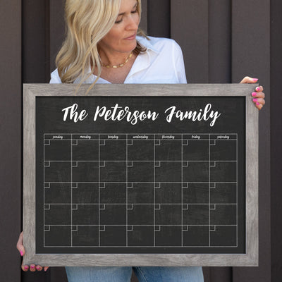 A woman stands against a dark wall, wearing a white blouse and pink nail polish, holding a framed chalkboard from CalendarGirlCompany titled new I - Medium Horizontal Chalkboard Template - 1 Magnet Set, ready for use with chalk ink markers.