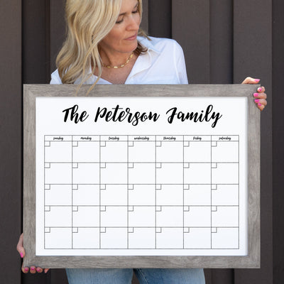 A person holds a 24 X 18 Framed Whiteboard Calendar by CalendarGirlCompany, featuring The Peterson Family at the top. The premium acrylic surface displays seven columns for days and five rows for weeks. Wearing a white shirt and necklace, they stand against a dark wall with colorful chalk ink markers ready.