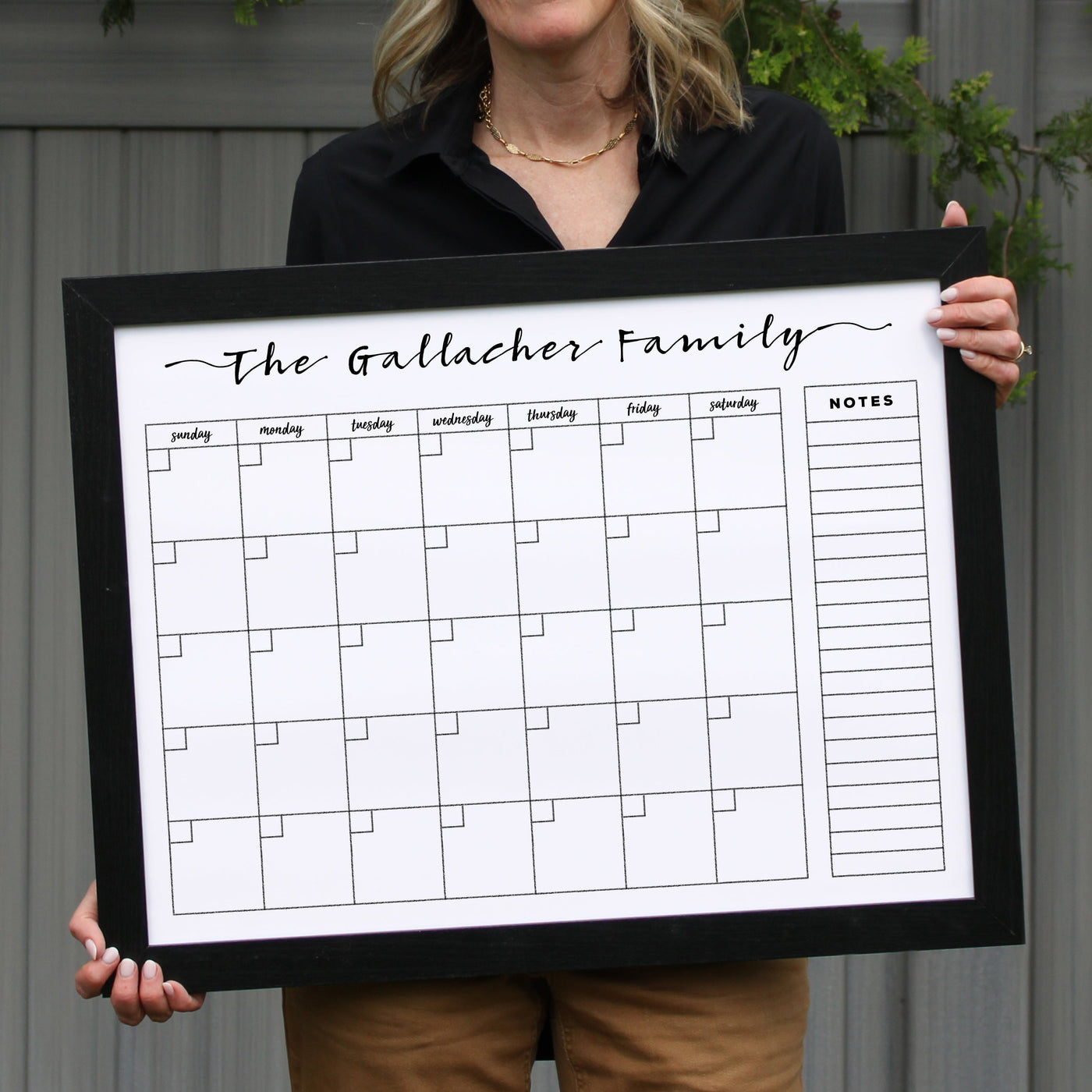 A person confidently stands against a gray wall holding the 24 X 18 Framed Whiteboard Calendar (#18203) by CalendarGirlCompany, labeled The Gallacher Family. The calendar includes a weekly grid and notes section, ideal for chalk ink markers. The individual is dressed in a black top and beige pants.