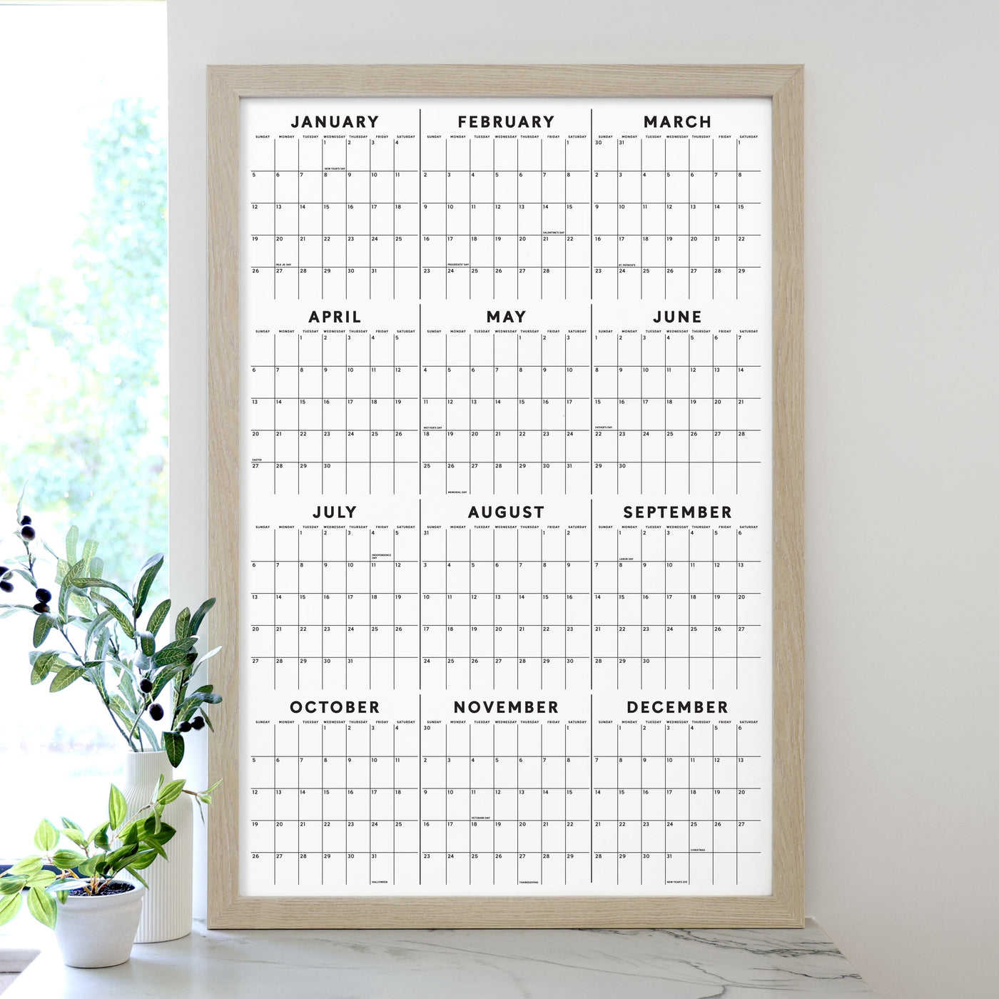 The Girl Friday 2025 Framed Yearly Calendar with Dates & Holidays, featuring a premium acrylic surface, hangs elegantly on the wall. Its minimalist design spans January to December and wet-erase markers rest nearby. A small green-leafed plant decorates the table beside it.