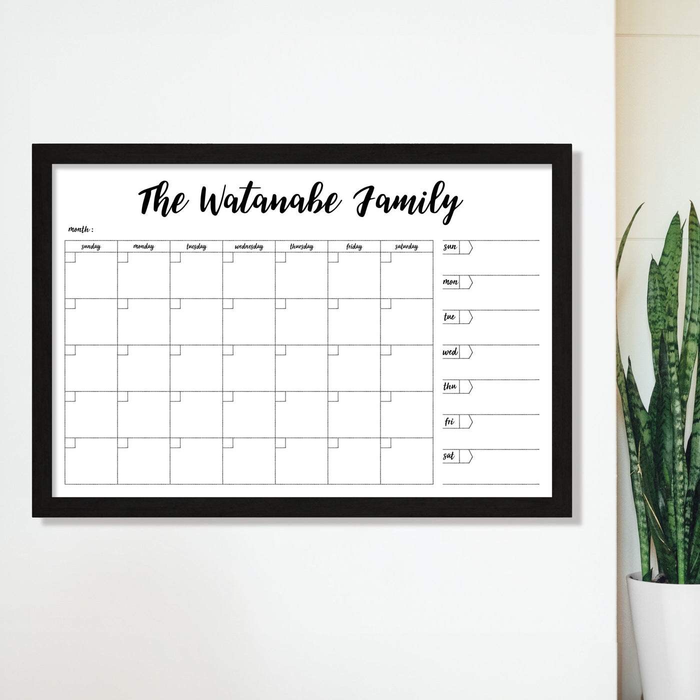 The CalendarGirlCompanys 36 x 24 Framed Whiteboard Calendar (#24126), labeled The Watanabe Family, features a premium acrylic surface with a weekly grid and note space. Its wall-mounted, black-framed, and accompanied by a lush plant on the right for a natural touch.