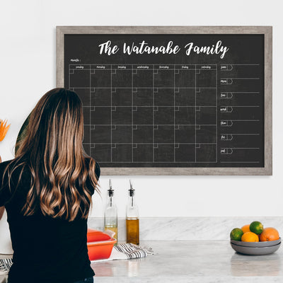 In a kitchen with marble counters hosting olive oil, vinegar, a red dish, and fruit, a woman writes on a CalendarGirlCompanys #24126 36 x 24 Framed Chalkboard Calendar using vibrant chalk ink markers. The calendar is labeled The Watanabe Family and mounted on a white wall in the background.