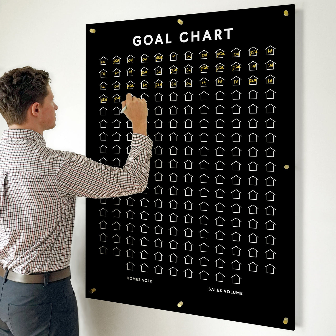 A man in a checkered shirt stands beside the Girl Friday Customizable Goal Chart, where he fills another outlined house icon. The black acrylic board prominently displays categories like HOMES SOLD and SALES VOLUME under the title GOAL CHART.
