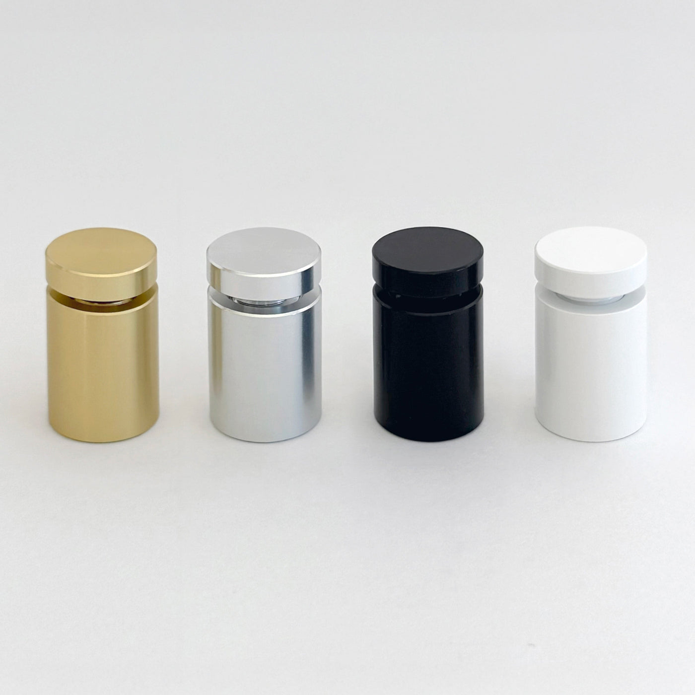 Four cylindrical containers are neatly lined up on a plain background, gleaming in metallic gold, silver, black, and white. Each has a matching round cap and pairs perfectly with accents from your Girl Friday Shops Clear Acrylic Monthly Wall Calendar | Black Text or a clear acrylic board.