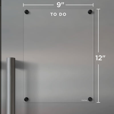 The Girl Friday Clear Acrylic Fridge To Do List, customizable with white text, measures 9x12 inches and mounts with four black corner standoffs. It features TO DO at the top and a left metallic handle suitable for wet-erase markers.
