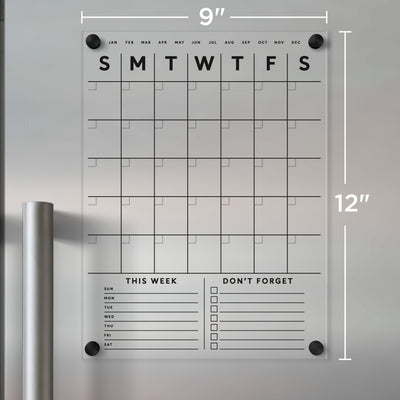 The Clear Acrylic Monthly Magnetic Fridge Calendar by Girl Friday Shop, measuring 9x12 inches, has a grid layout with months, This Week, and Dont Forget sections. With black text and small black mounting circles in the corners, its perfect for dry-erase markers.