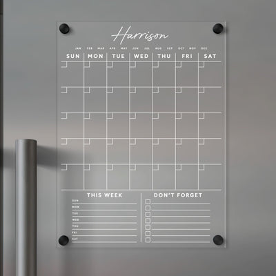 The Clear Acrylic Monthly Fridge Calendar from Girl Friday features an elegant monthly grid for daily entries. It includes customizable This Week and Dont Forget sections at the bottom, ideal for wet-erase markers, and personalization with your family name in white text.