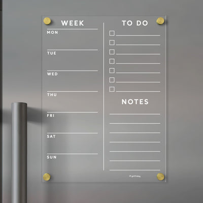 The Girl Friday Acrylic Fridge Planner, mounted with gold standoffs, offers customizable daily sections, a to-do list with checkboxes, and a notes area. Ideal for dry-erase markers, it features a slight metallic hint on the left side.