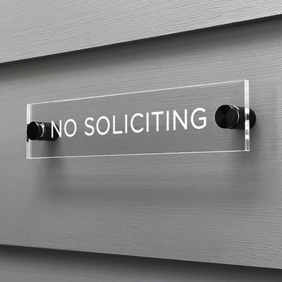 The Girl Friday Shops No Soliciting acrylic sign, featuring bold white text, is securely mounted on a gray wall using sleek black hardware.