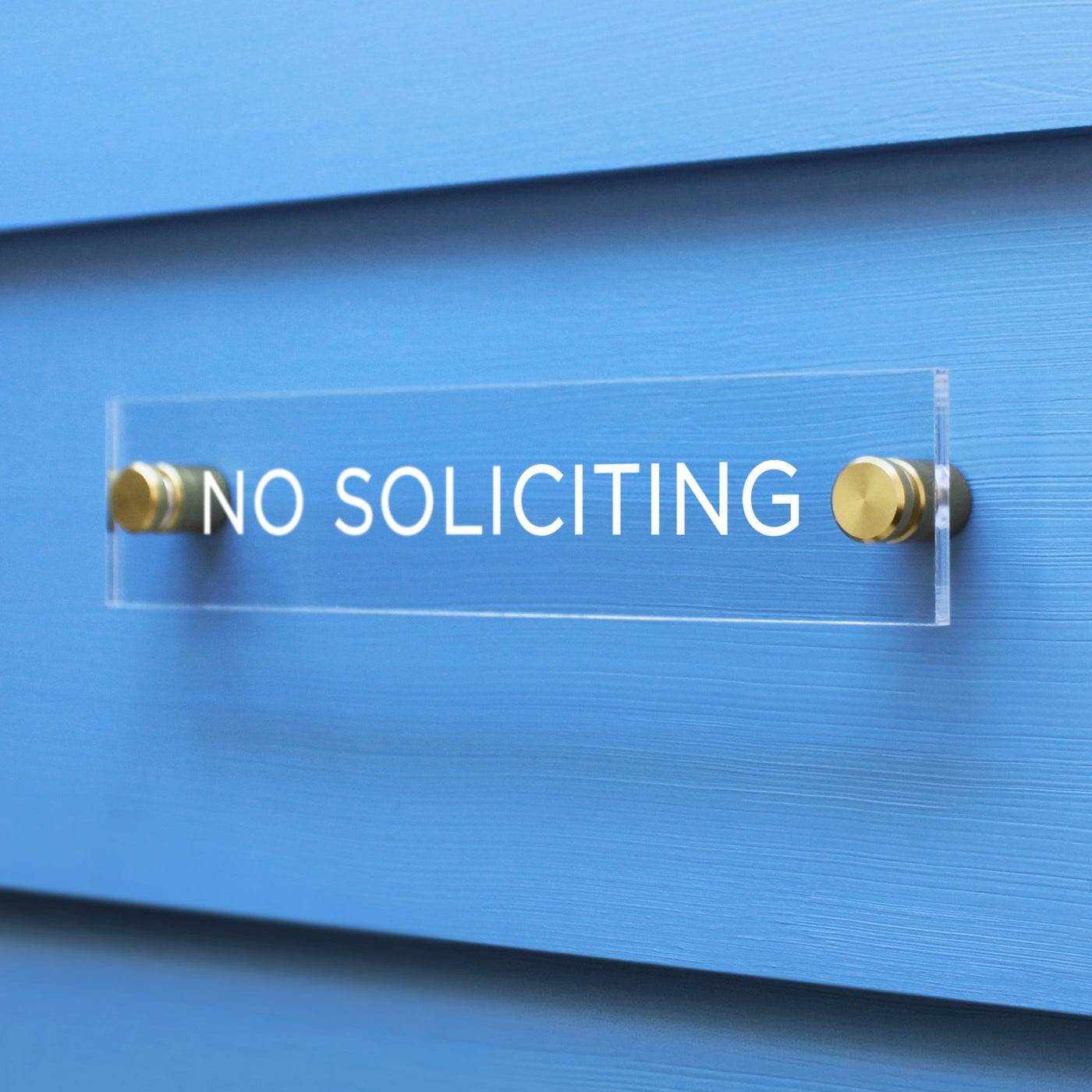 The Girl Friday Shops No Soliciting Acrylic sign, featuring white text, is mounted on a bright blue wall with two gold fasteners.