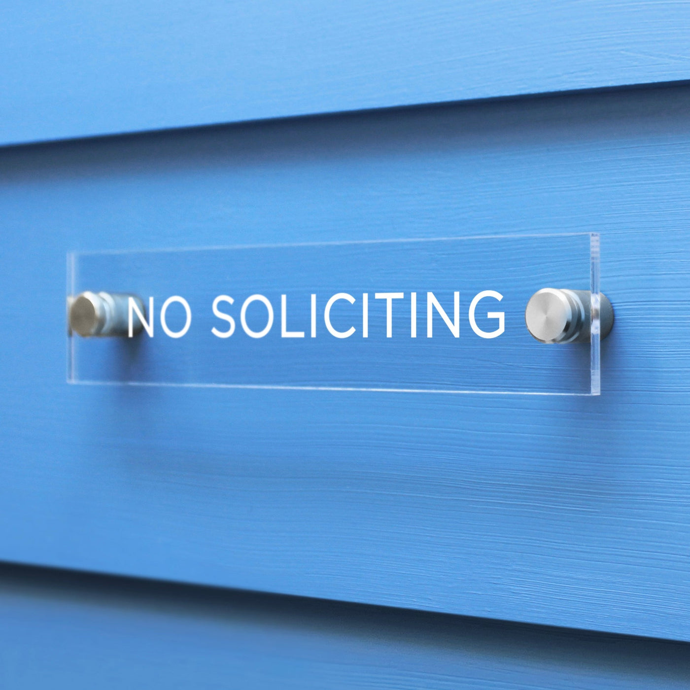 A No Soliciting Acrylic sign by Girl Friday Shop is securely mounted on a light blue wall with metallic bolts.