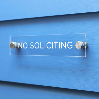 A No Soliciting Acrylic sign by Girl Friday Shop is securely mounted on a light blue wall with metallic bolts.