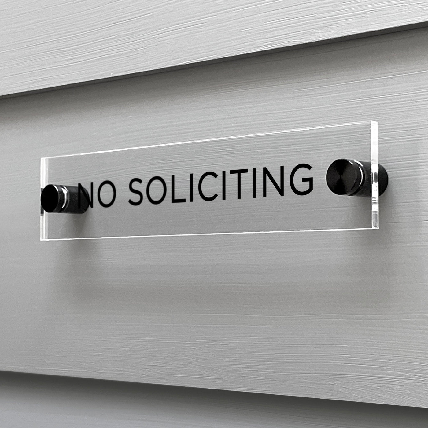 A sleek Girl Friday Shop No Soliciting Acrylic Sign, featuring bold black text, is firmly mounted on a gray wall with two screws. This sign can be personalized to suit your needs while keeping its striking clarity.
