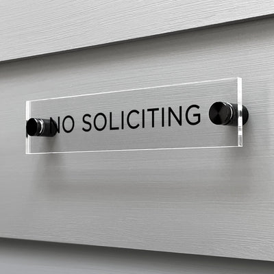 A sleek Girl Friday Shop No Soliciting Acrylic Sign, featuring bold black text, is firmly mounted on a gray wall with two screws. This sign can be personalized to suit your needs while keeping its striking clarity.