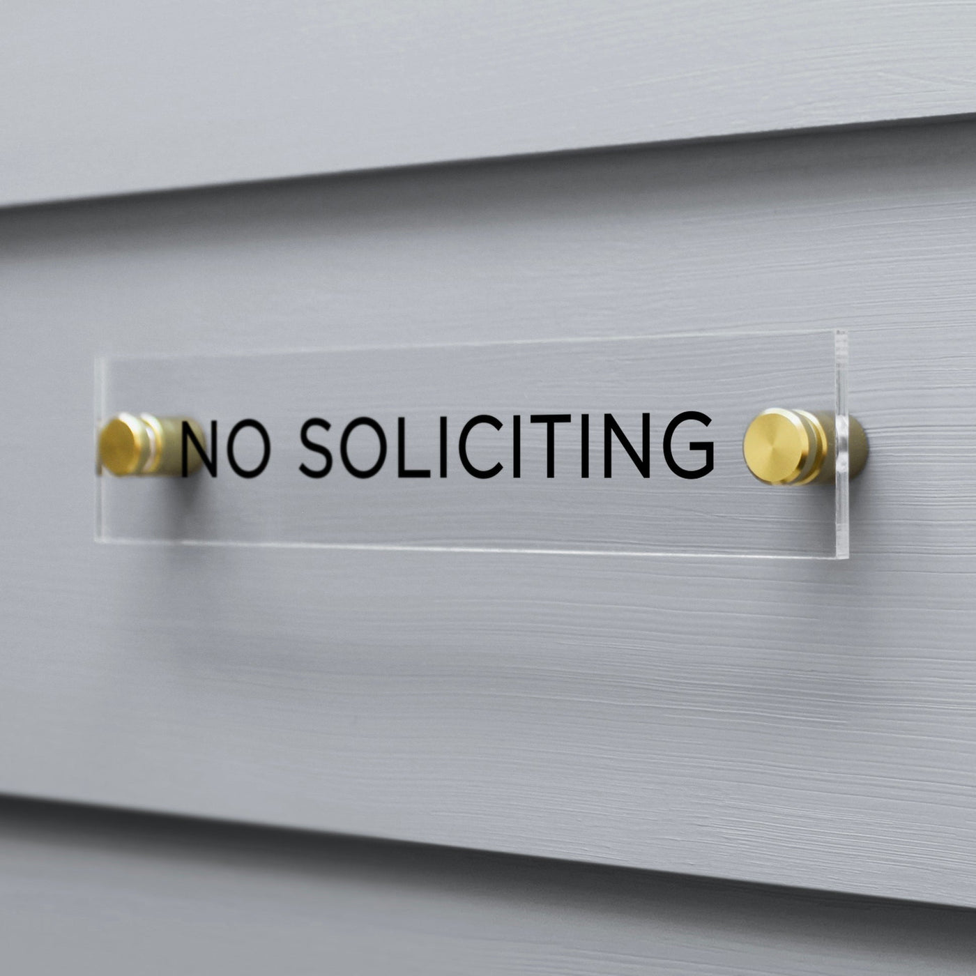 The Girl Friday Shops No Soliciting Acrylic Sign features bold black text and is mounted with gold screws against gray siding, offering a clear and stylish message.