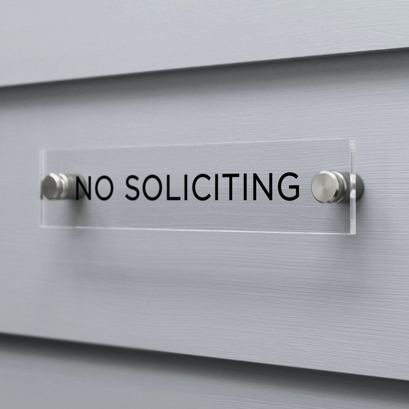 The No Soliciting Acrylic sign by Girl Friday Shop, featuring black text, is mounted on a gray wall using two metal fasteners.