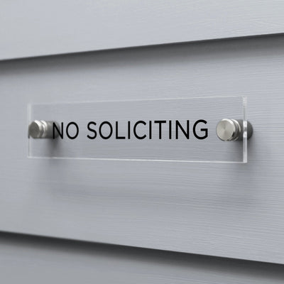 The No Soliciting Acrylic sign by Girl Friday Shop, featuring black text, is mounted on a gray wall using two metal fasteners.