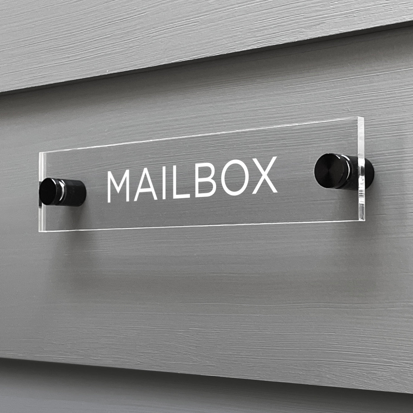 The Mailbox Acrylic Sign by Girl Friday Shop, featuring bold white letters, is custom-designed and elegantly mounted on a gray wooden wall with two cylindrical metal posts.