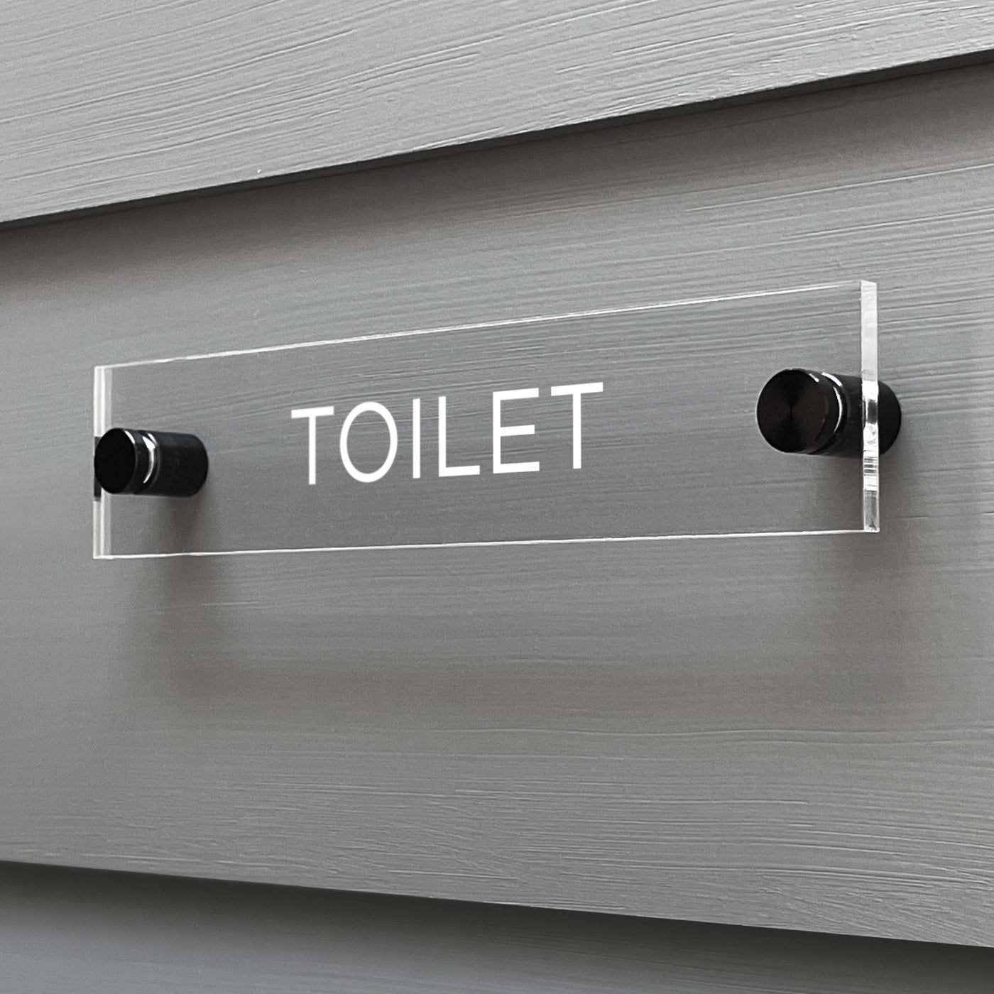 The Girl Friday Shop Toilet Acrylic Sign, with TOILET in bold white letters, is a sleek rectangular design mounted on a gray wall and securely held by two black pegs.