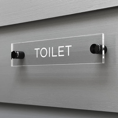 The Girl Friday Shop Toilet Acrylic Sign, with TOILET in bold white letters, is a sleek rectangular design mounted on a gray wall and securely held by two black pegs.