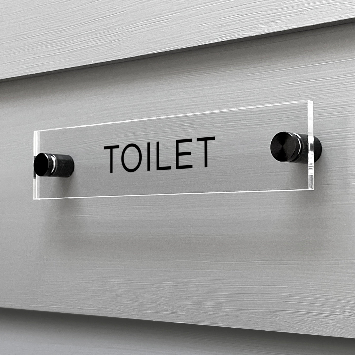 The Girl Friday Shops Toilet Acrylic Sign, featuring a custom TOILET design, adds a sleek touch when mounted on a gray wall.