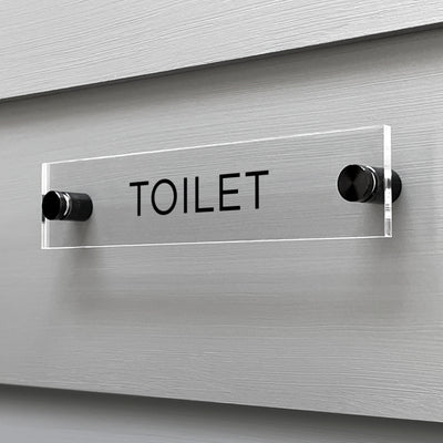 The Girl Friday Shops Toilet Acrylic Sign, featuring a custom TOILET design, adds a sleek touch when mounted on a gray wall.