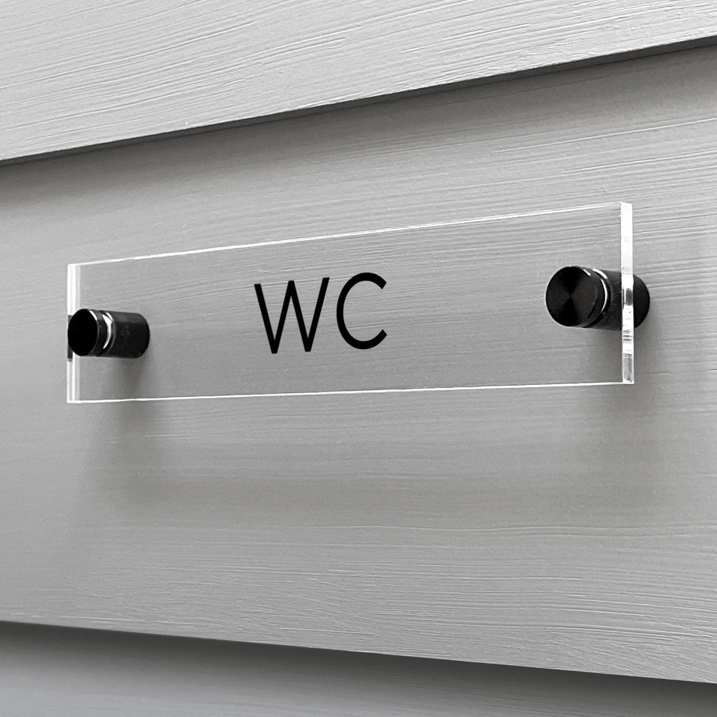 The Girl Friday Shops WC Acrylic Sign, featuring bold black letters, is mounted on a light gray wall with two cylindrical black supports, creating a sleek contrast against the textured horizontal panel for modern elegance.