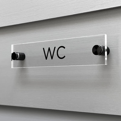 The Girl Friday Shops WC Acrylic Sign, featuring bold black letters, is mounted on a light gray wall with two cylindrical black supports, creating a sleek contrast against the textured horizontal panel for modern elegance.